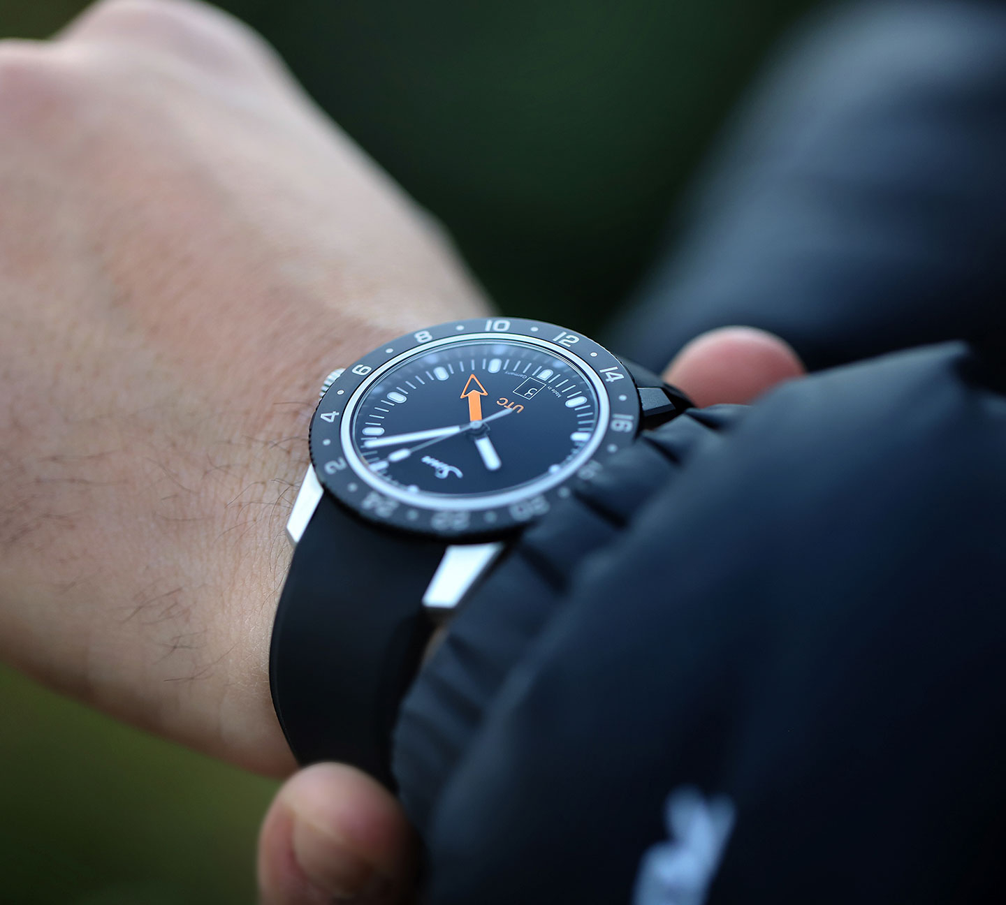 Sinn 105 St Sa UTC mit schwarzem Zifferblatt, orangefarbenem UTC-Pfeilzeiger und schwarzem Kautschukarmband am Handgelenk.