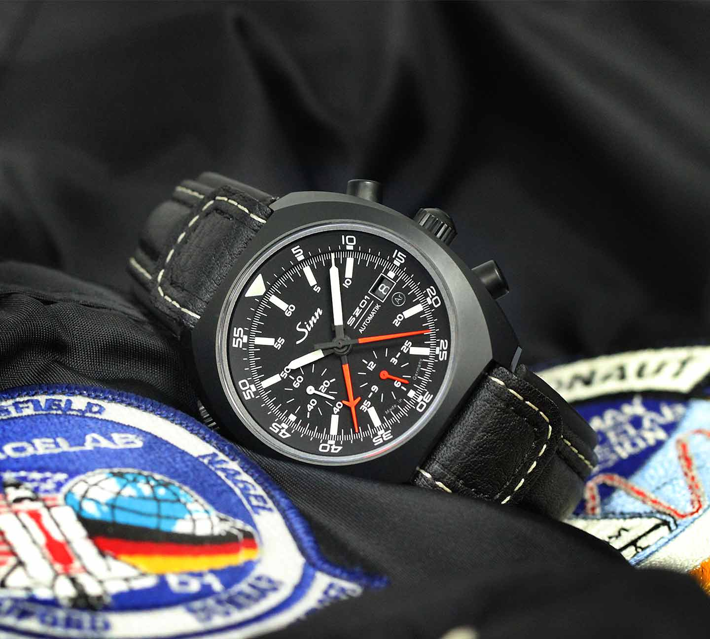 Sinn 140 St S Automatik-Chronograph mit schwarzem Gehäuse, schwarzem Zifferblatt, roten und weißen Zeigern und Lederarmband, platziert auf einer Astronautenjacke mit Missionsabzeichen.