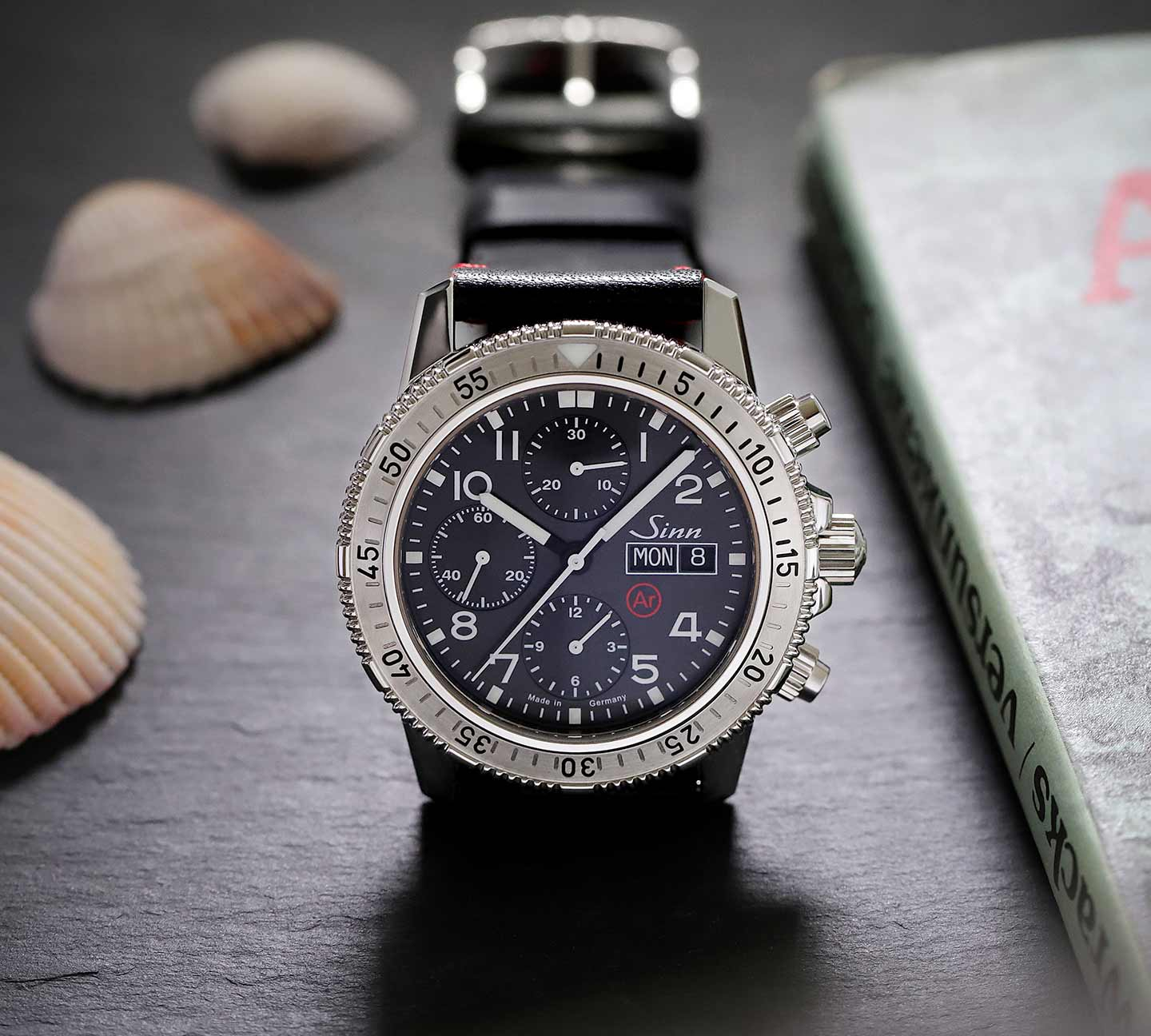 Sinn 206 St Ar mit schwarzem Zifferblatt und Lederband, dekorativ inszeniert mit Muscheln und Buch.