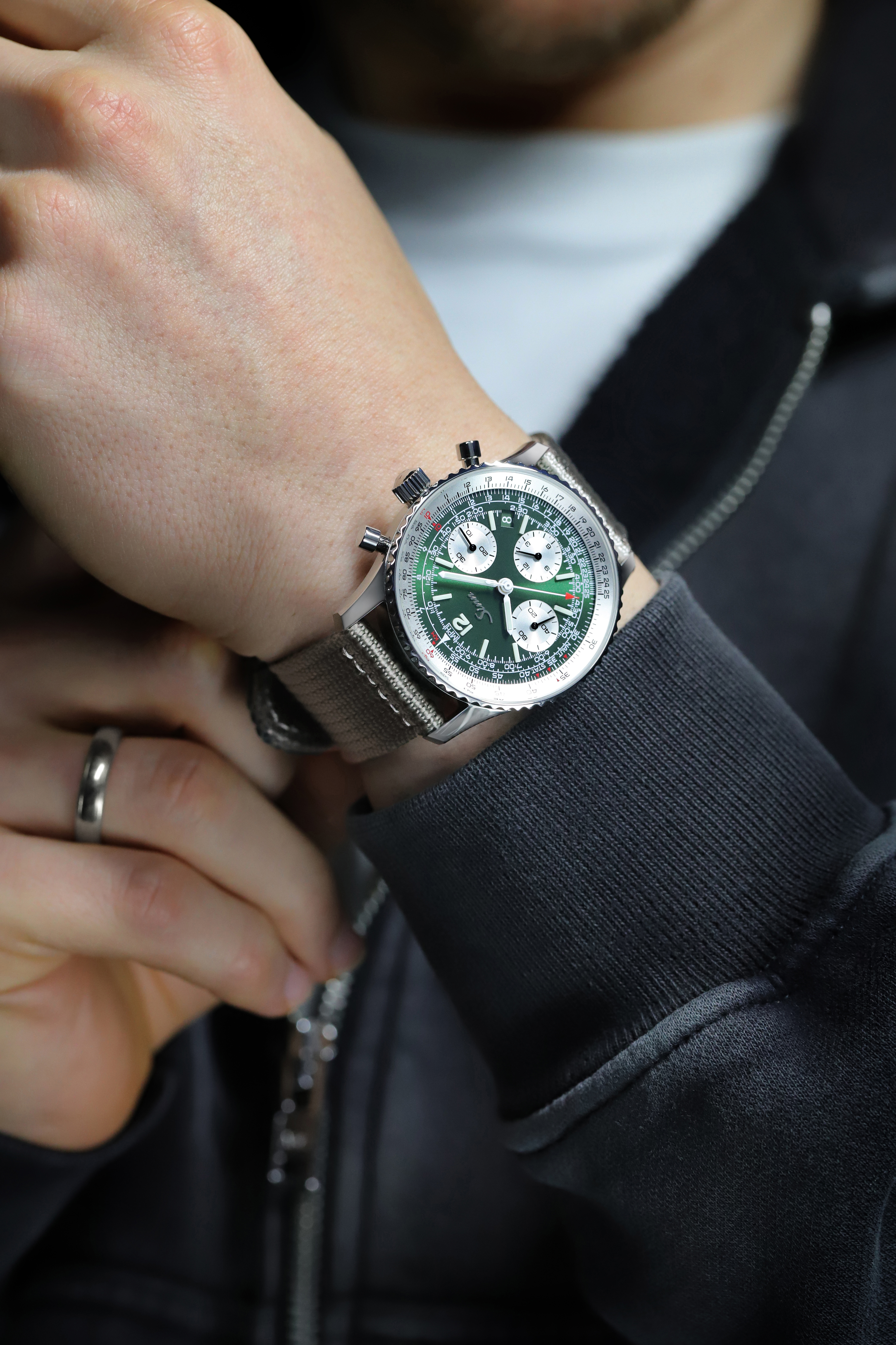 Sinn chronograph with green dial, slide rule bezel, and grey textile strap worn on a wrist.