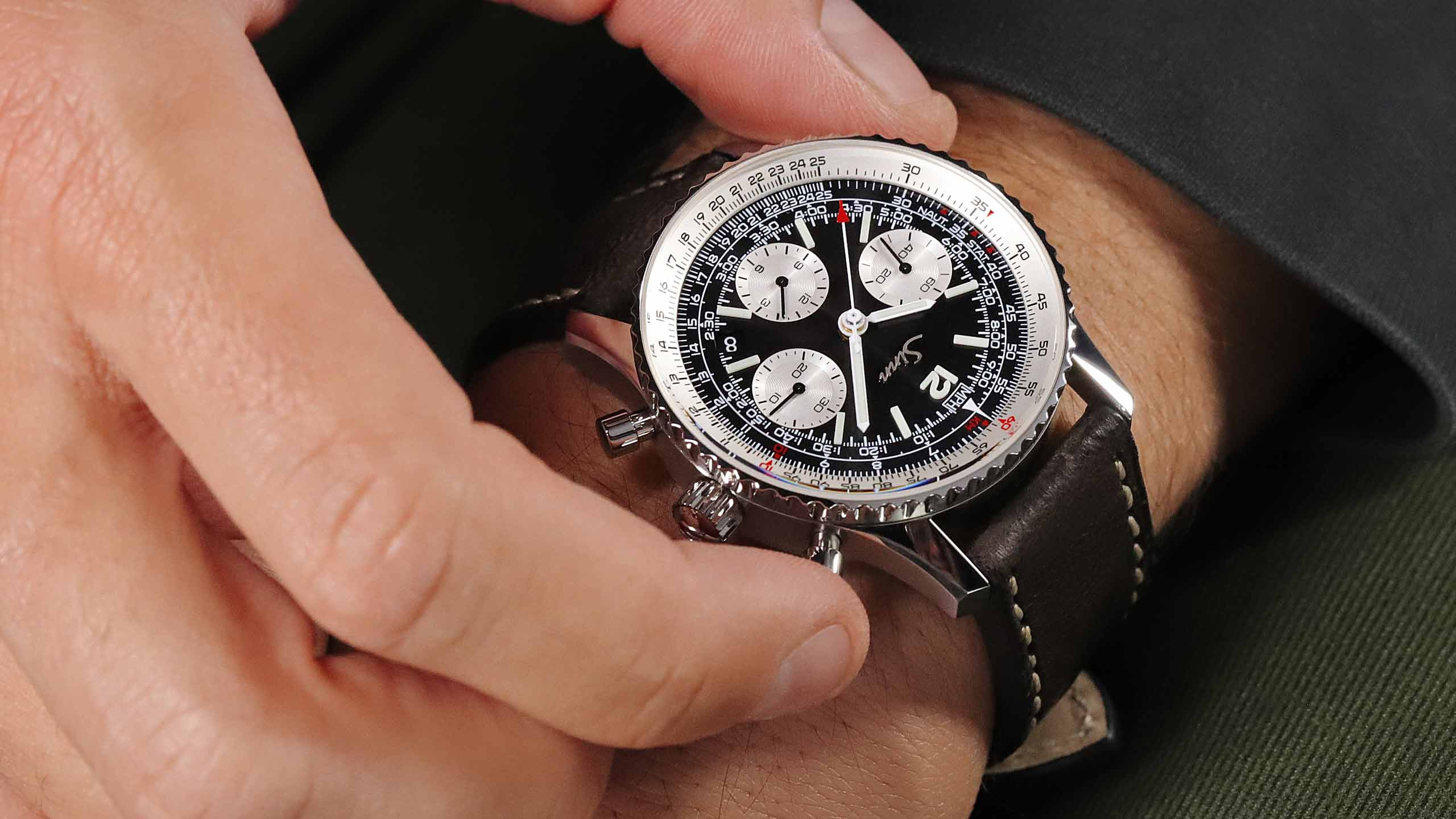                       Close-up of a Sinn chronograph with black dial and white subdials on a leather strap, being adjusted on a wrist
                  