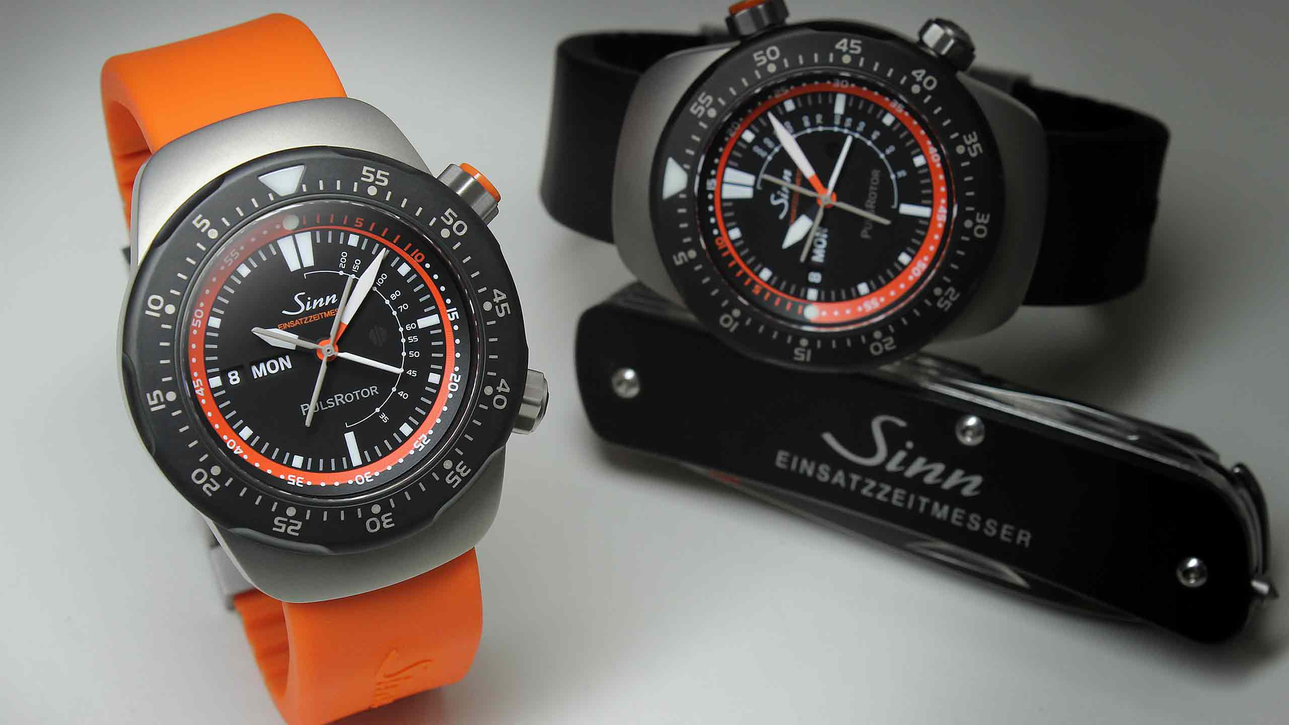                       Zwei Sinn EZM 12 Uhren, eine mit orangefarbenem Silikonband, eine mit schwarzem Band, daneben ein schwarzes Sinn-Einsatzzeitmesser-Taschenmesser.
                  