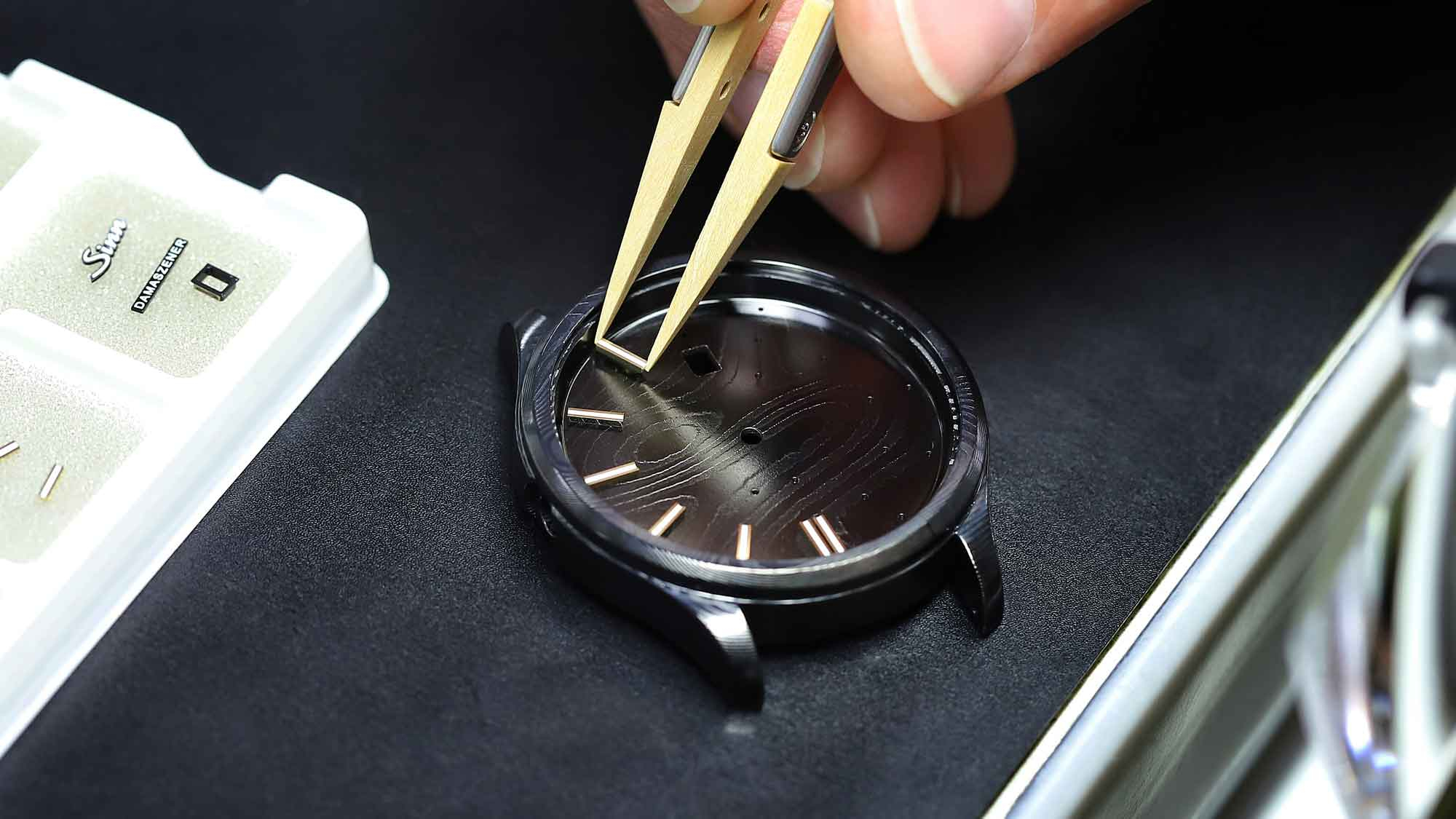 Watchmaker applies gold appliques to a Damascus steel Sinn watch dial with tweezers.