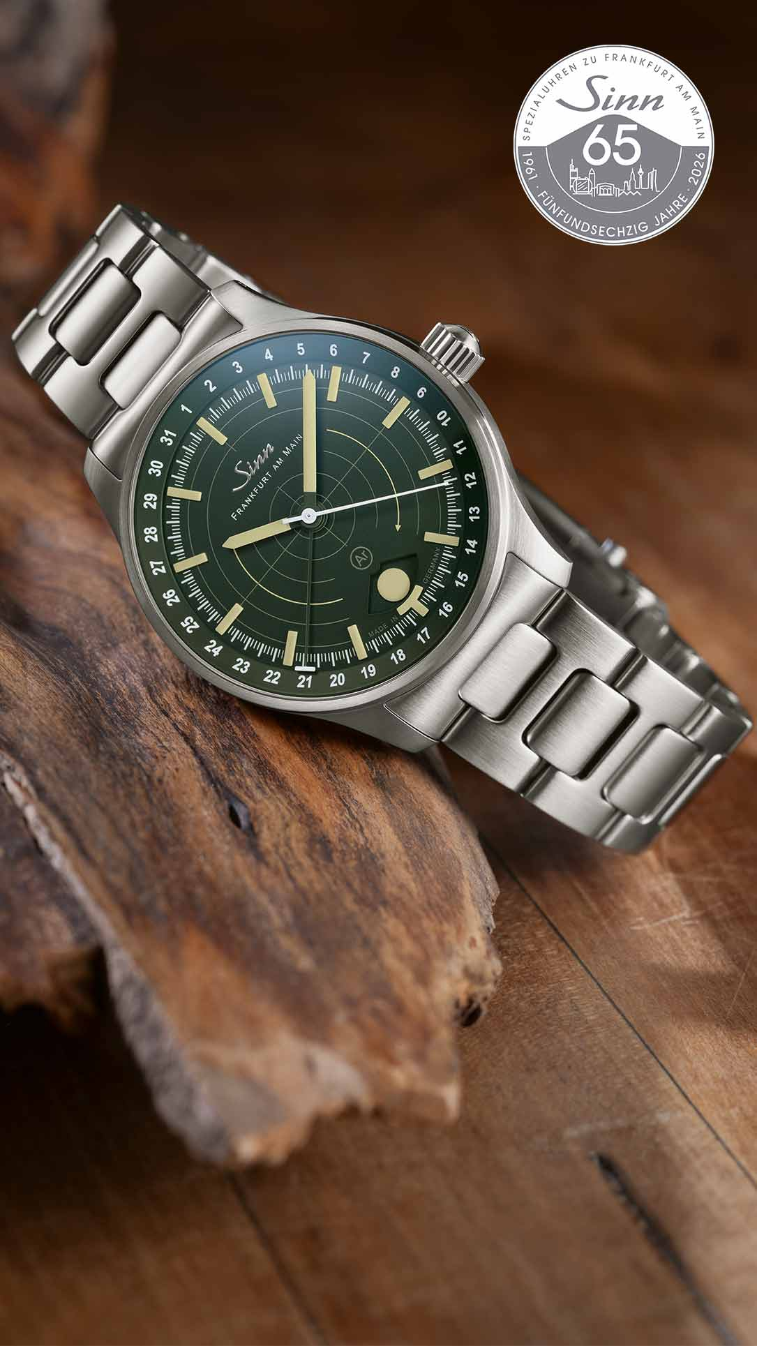 Stainless steel wristwatch with a green dial and metal bracelet, placed on a rustic wooden surface; anniversary logo on the right side.