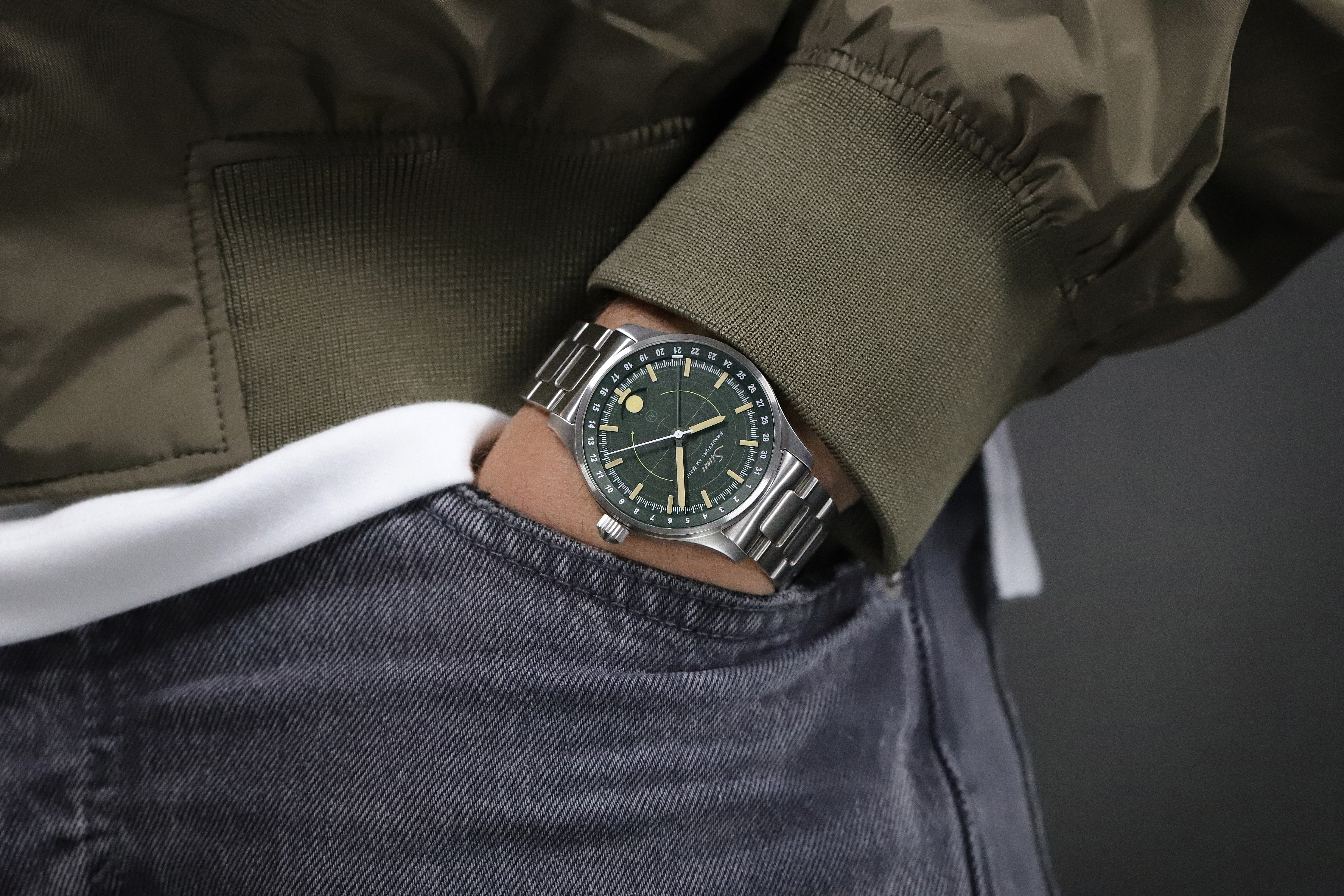 Armbanduhr mit grünem Zifferblatt und Metallarmband am Handgelenk, kombiniert mit einer Jacke und Jeans.