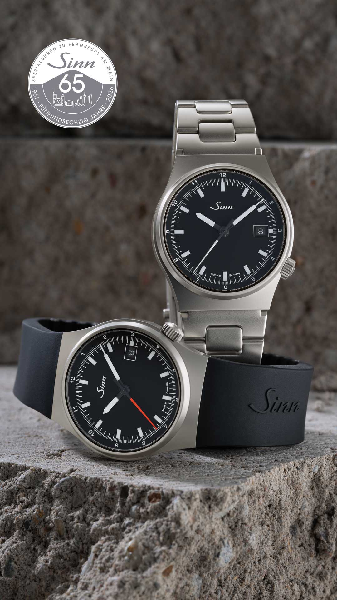 Two stainless steel wristwatches with black dials—one with a metal bracelet and one with a black silicone strap—arranged on a rough stone surface; anniversary logo on the right.