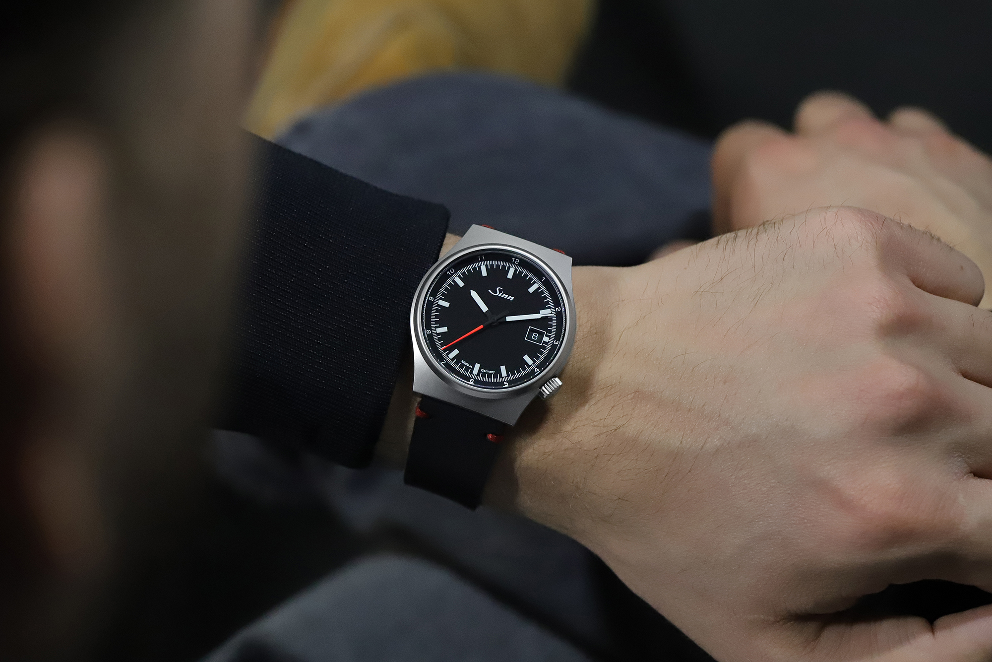 Montre-bracelet avec cadran noir et trotteuse rouge portée au poignet, associée à des vêtements sombres.