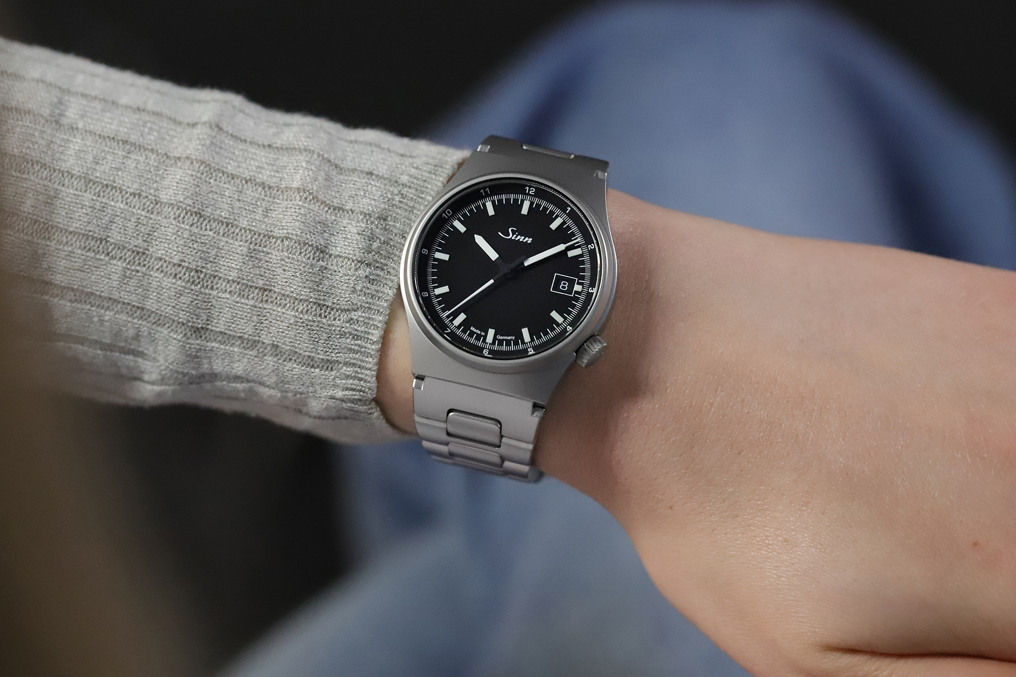 Wristwatch with a black dial and metal bracelet worn on the wrist, paired with a light knit sweater.