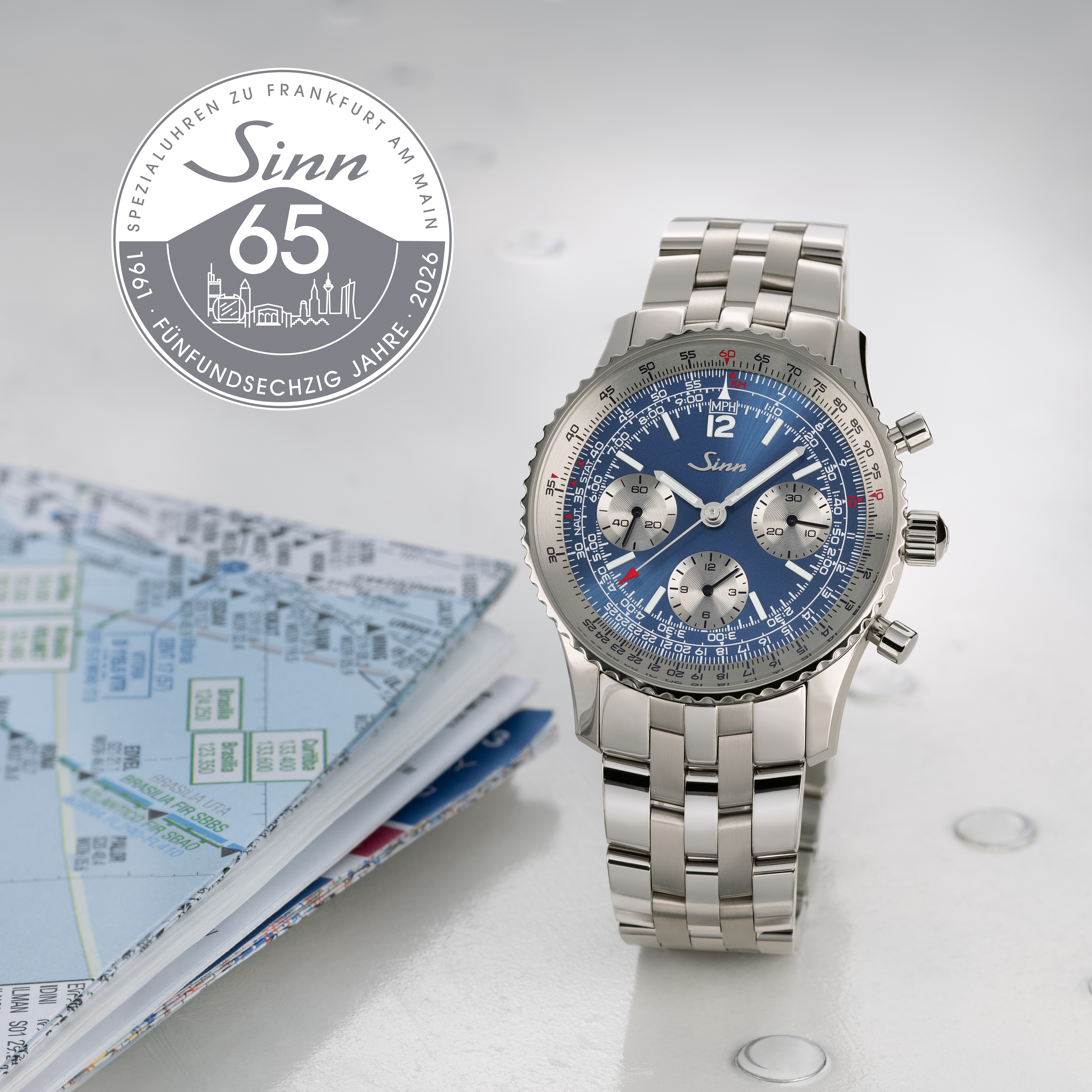 Sinn chronograph with blue dial and stainless steel bracelet next to an open notebook with aviation maps, featuring the Sinn 65th anniversary logo.