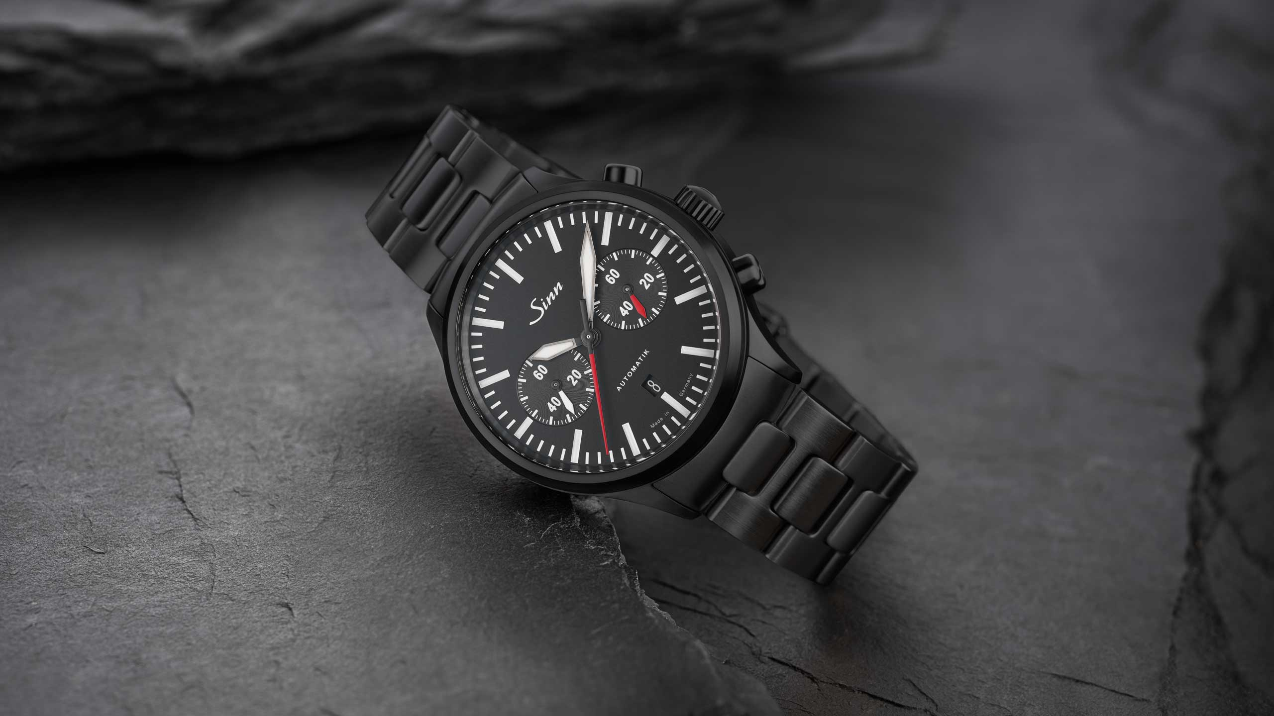                       Chronographe noir en acier inoxydable avec cadran sombre et bracelet métallique, posé sur une surface en pierre texturée
                  