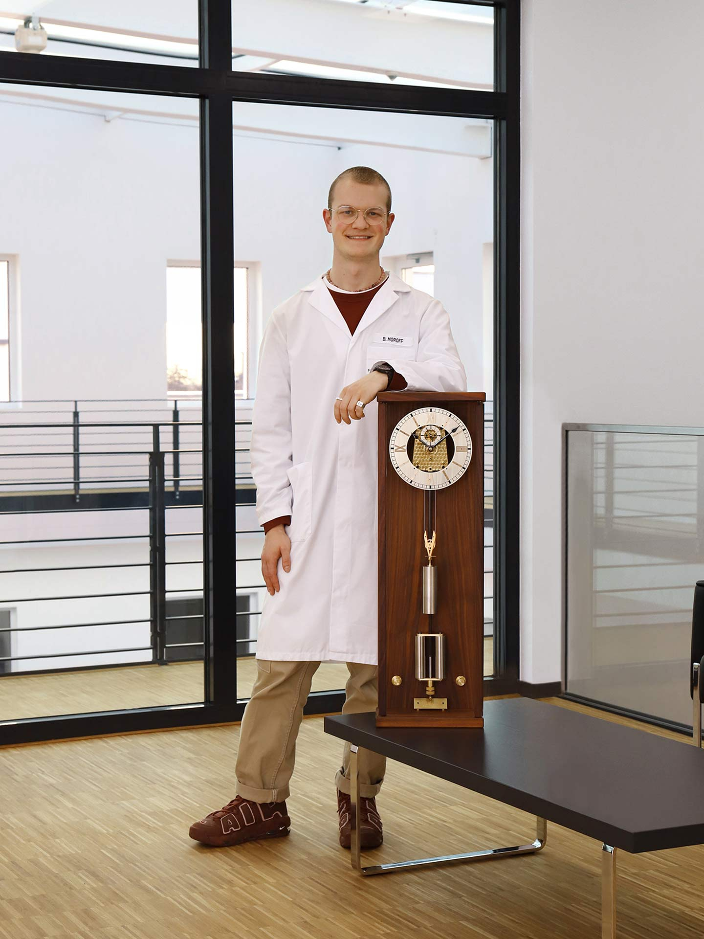 An employee of Sinn Spezialuhren wearing a white lab coat stands next to a mechanical clock placed on a table in a modern interior with large windows.