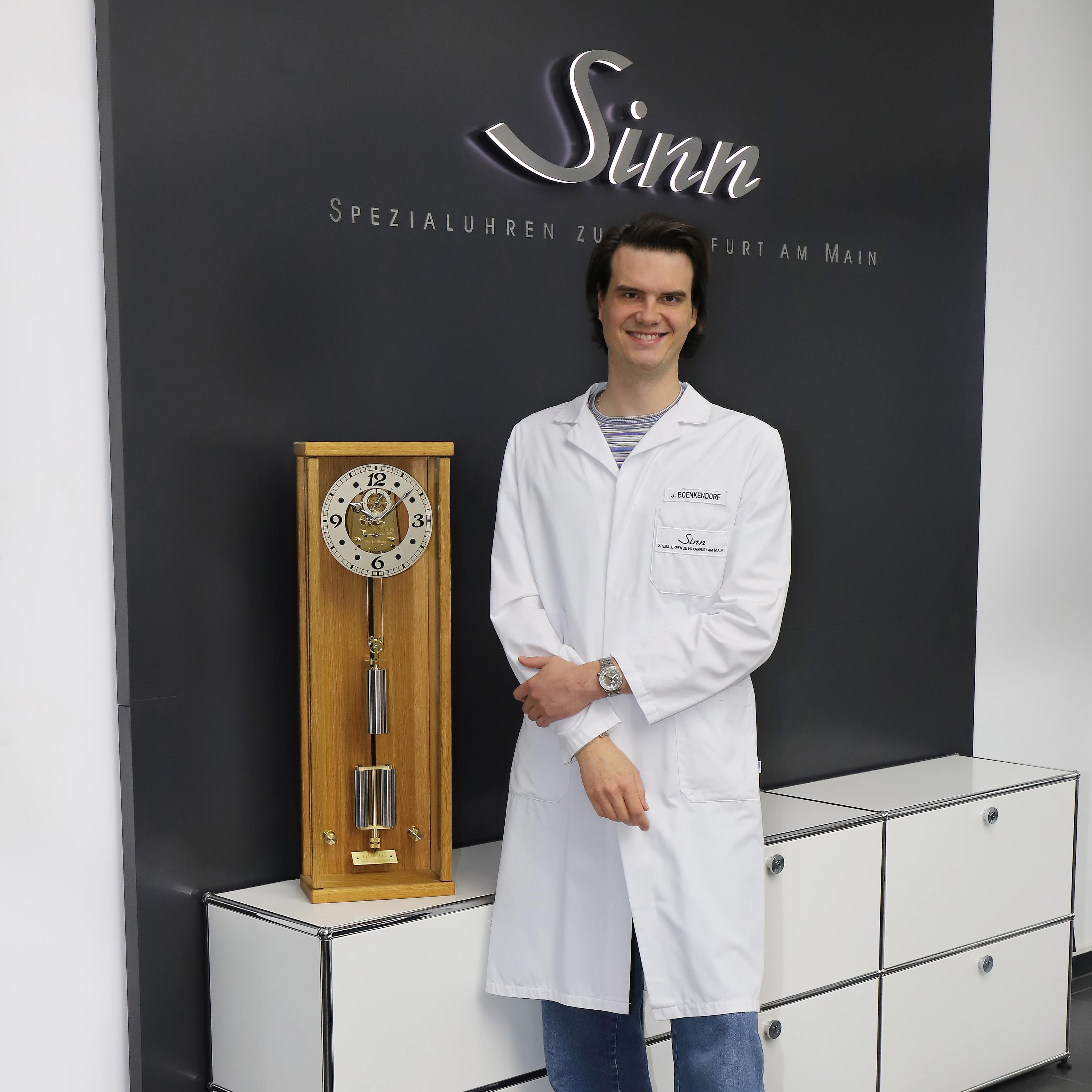 Watchmaker in a white lab coat standing next to a wooden mechanical pendulum clock in front of a wall with the Sinn Spezialuhren logo.