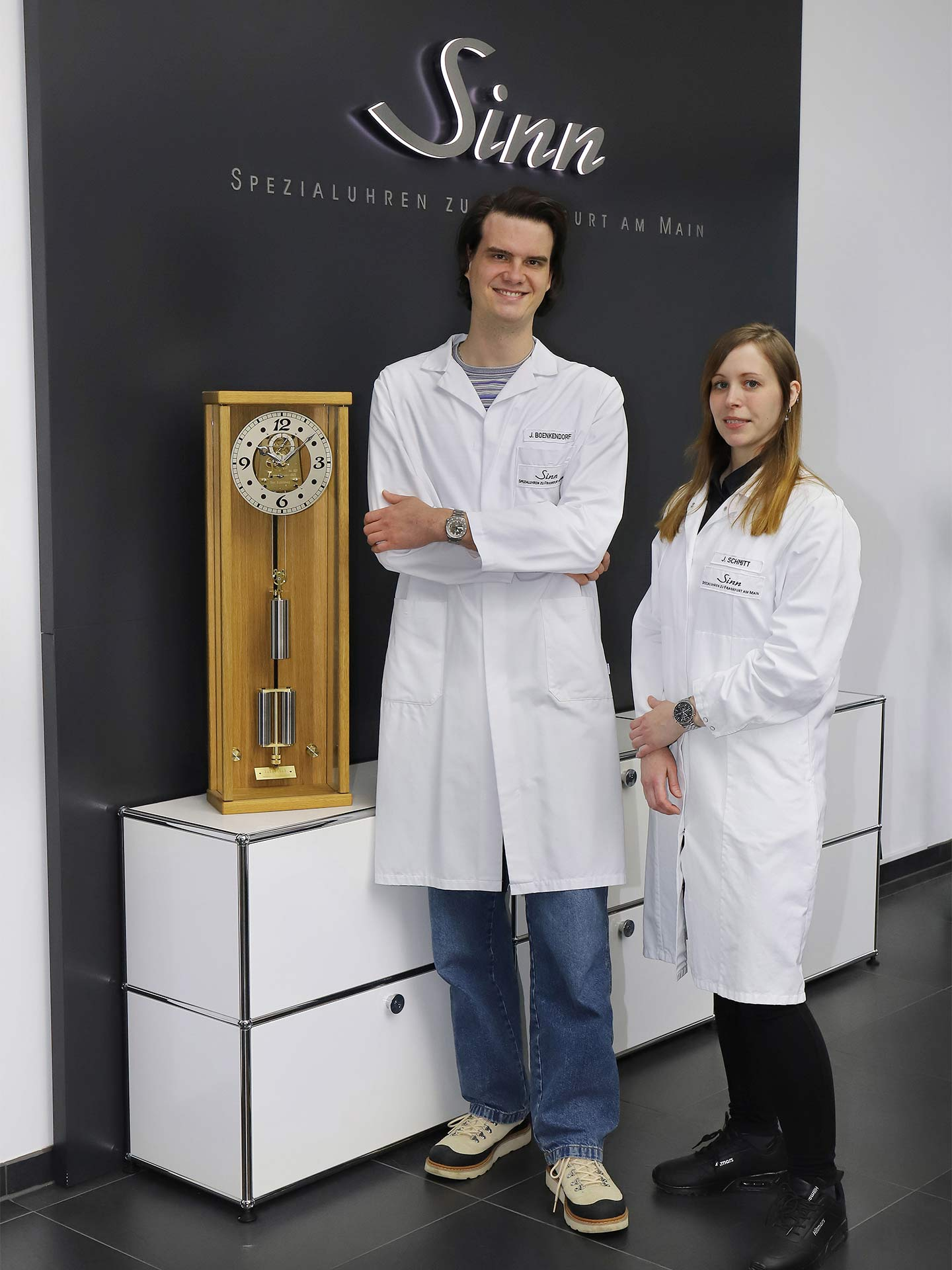 Two employees of Sinn Spezialuhren wearing white lab coats stand next to a large mechanical grandfather clock in front of a wall with the Sinn logo.