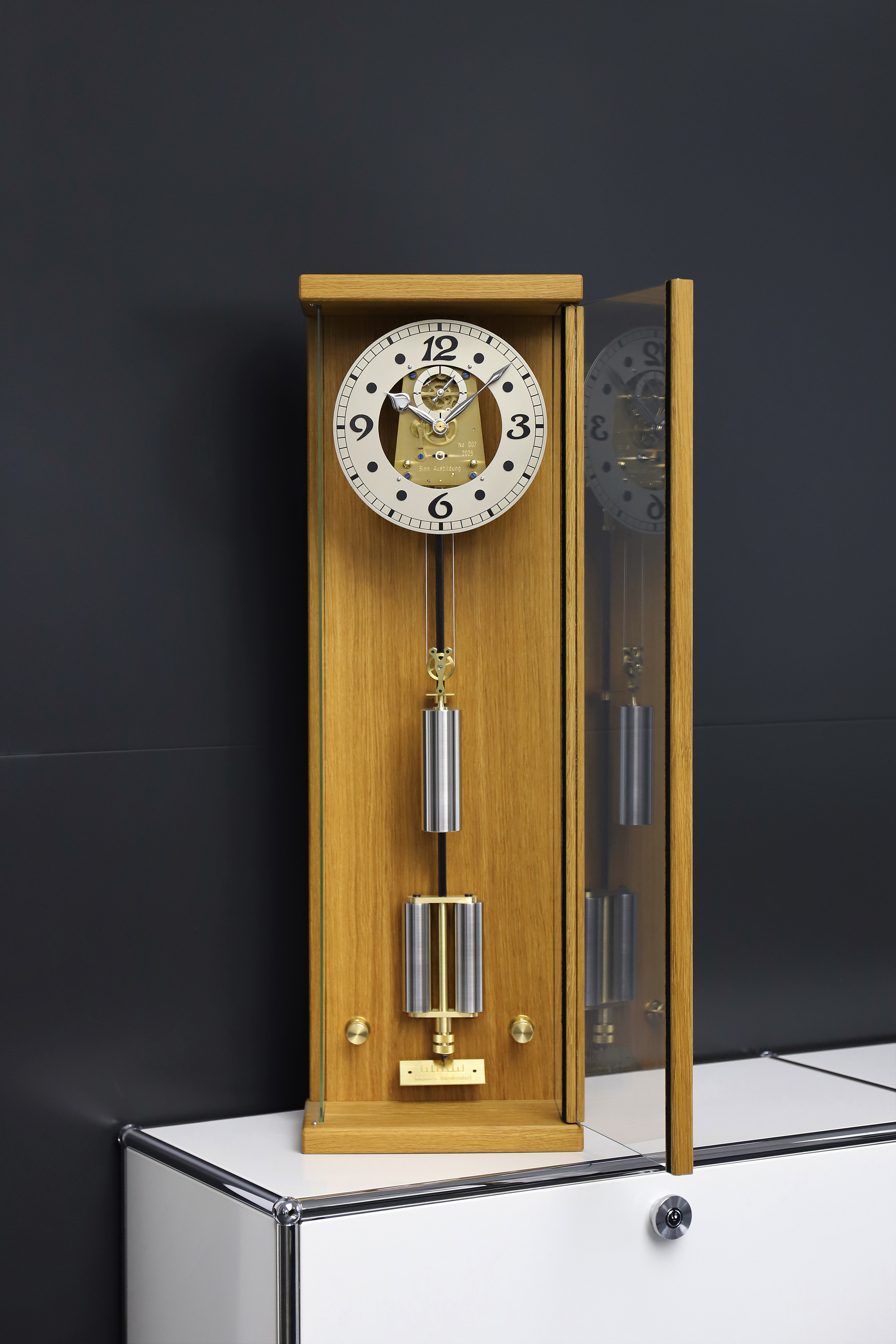 Open wooden mechanical clock with visible pendulum and dial, placed on a sideboard against a dark background.