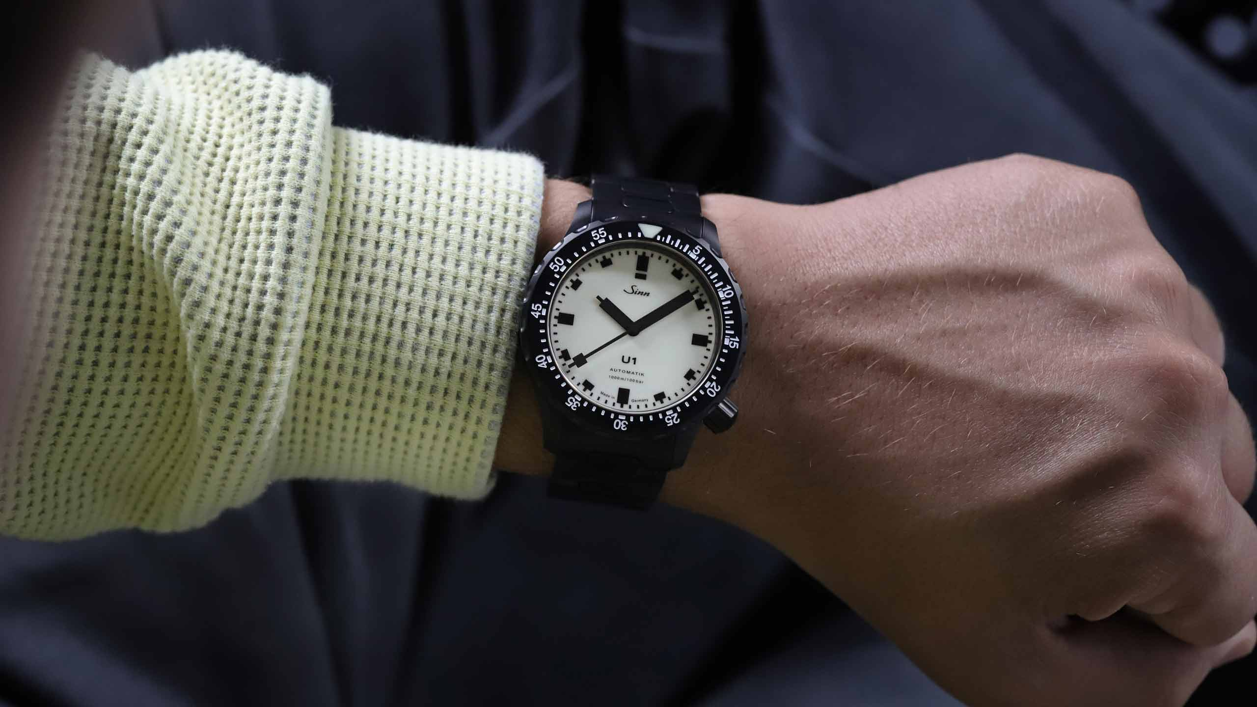                       Wristwatch with a black case and white dial on a person’s wrist, worn with a light-colored textured long-sleeve shirt.
                  