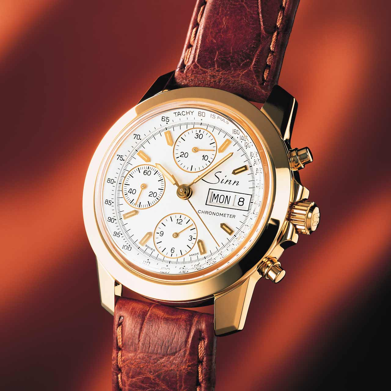 Sinn 2200 chronometer in gold with white dial, gold indices and brown leather strap, set against a warm-toned background