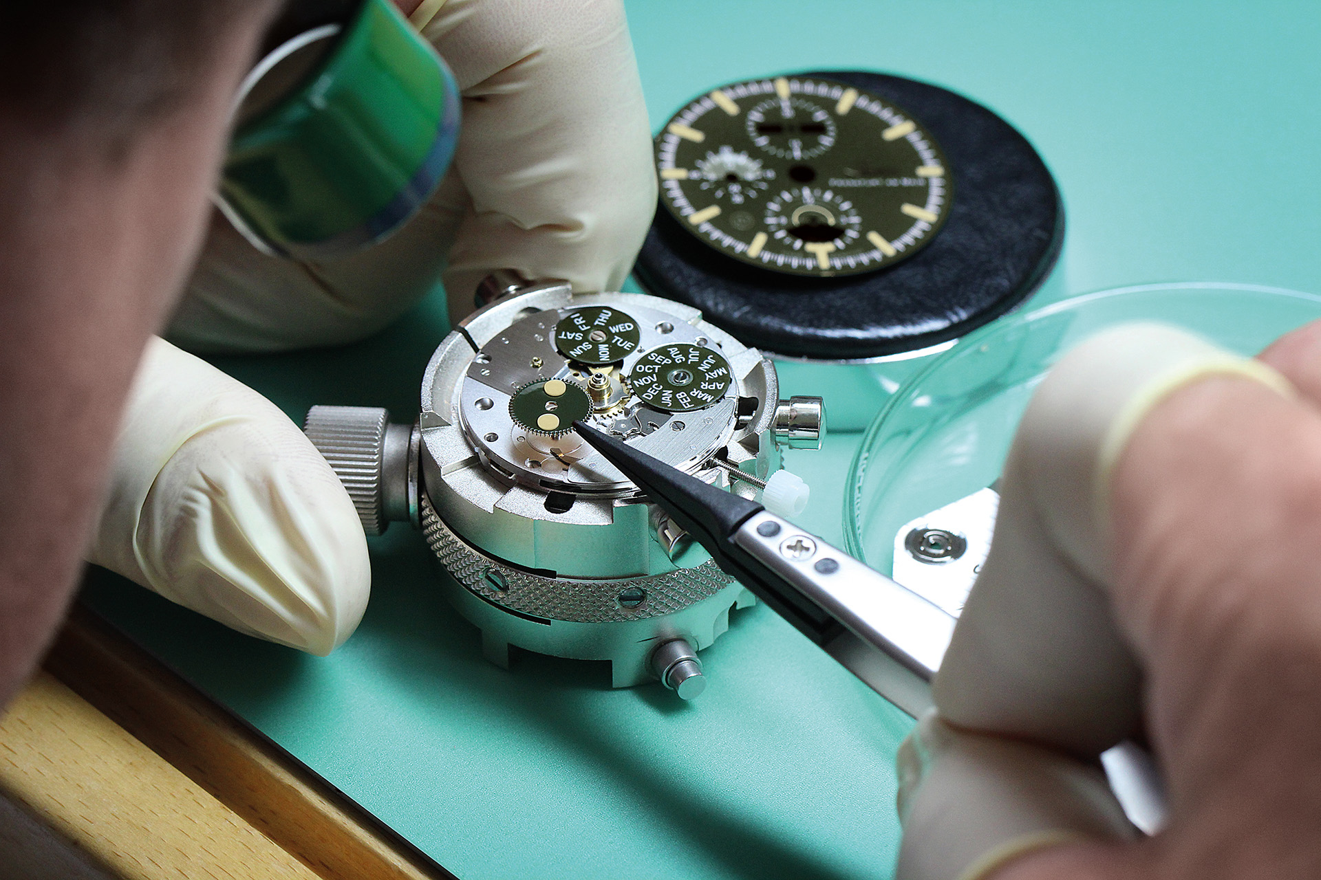 Watchmaker wearing gloves uses tweezers to install a moon phase disc in the movement of a Sinn wristwatch; the dial rests on the workbench in the background.
