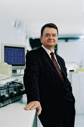 Lothar Schmidt im Anzug und Krawatte, steht vor einem Arbeitsplatz mit Monitor und Messgeräten.